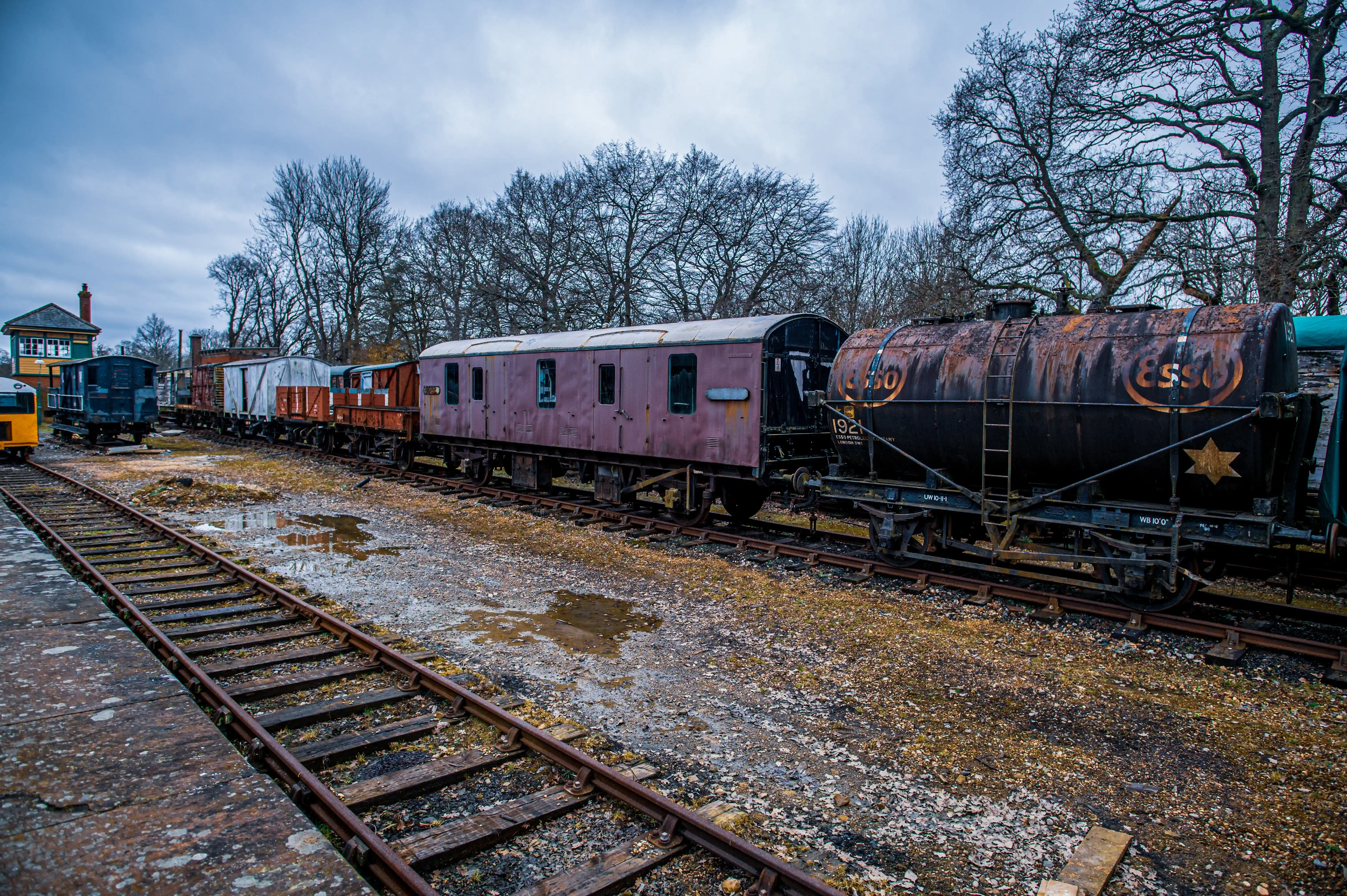 ESSO tanker and assorted vintage freight wagons standing in a heritage railway yard with gravel ground and winter trees.
